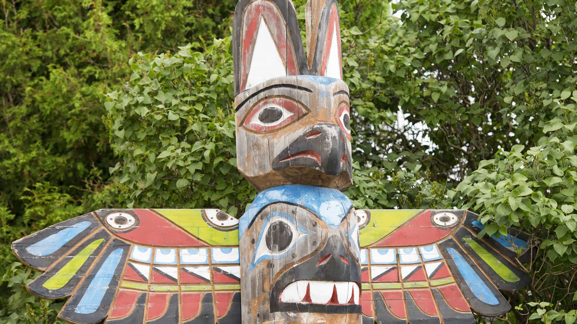 A detailed view of a traditional totem pole in Alaska, carved and painted with intricate Native American designs. The colors are faded, but you can still see red, white, black, blue, and green on the pole's figures. The totem pole features a stylized animal, possibly a bird or wolf, with a second face carved into its chest. The background is a dense, lush green forest. The image represents the rich cultural history and artistry of the Alaskan region.