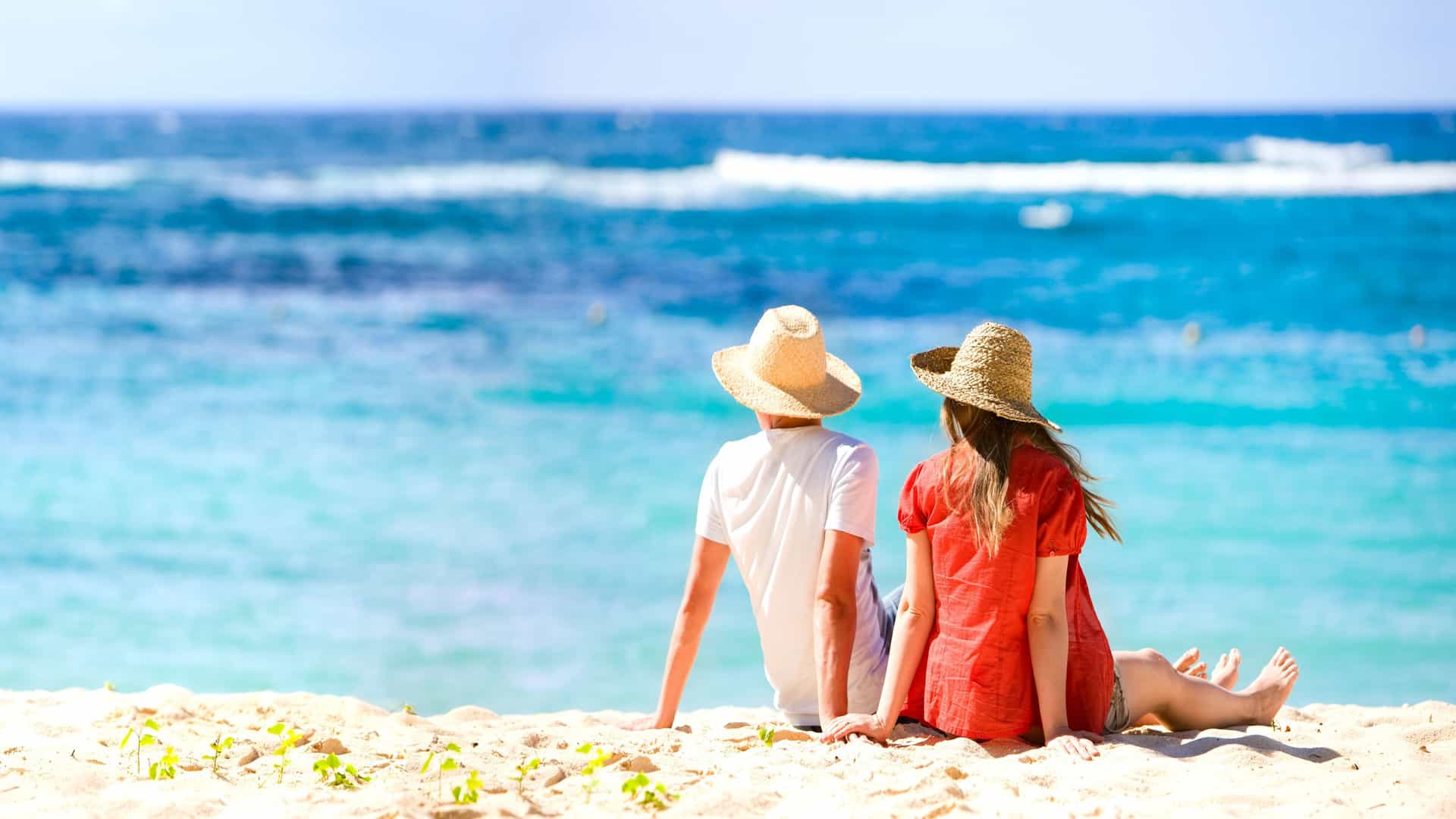 Two people sitting on the Caribbean beach, wearing hats, enjoying the ocean view.