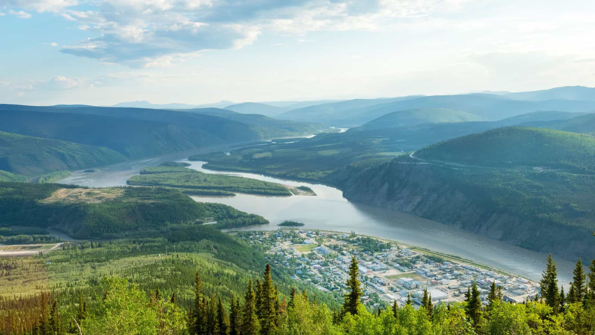 A stunning, high-angle view from Midnight Dome overlooking historic Dawson City, Yukon, nestled at the confluence of the Klondike and Yukon rivers amidst rolling green mountains.