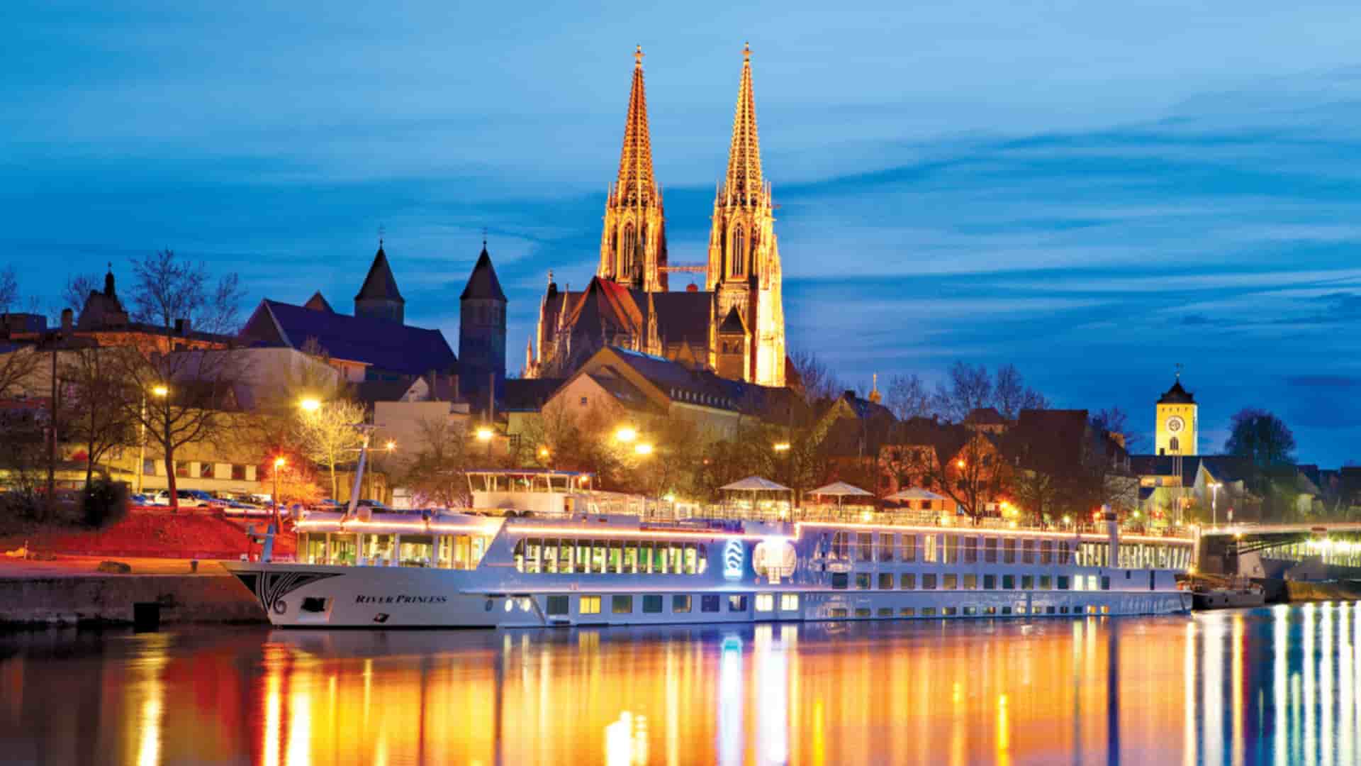 Uniworld's River Princess cruise ship docked on the Danube River in Regensburg, Germany, with the historic St. Peter's Cathedral illuminated against the night sky.
