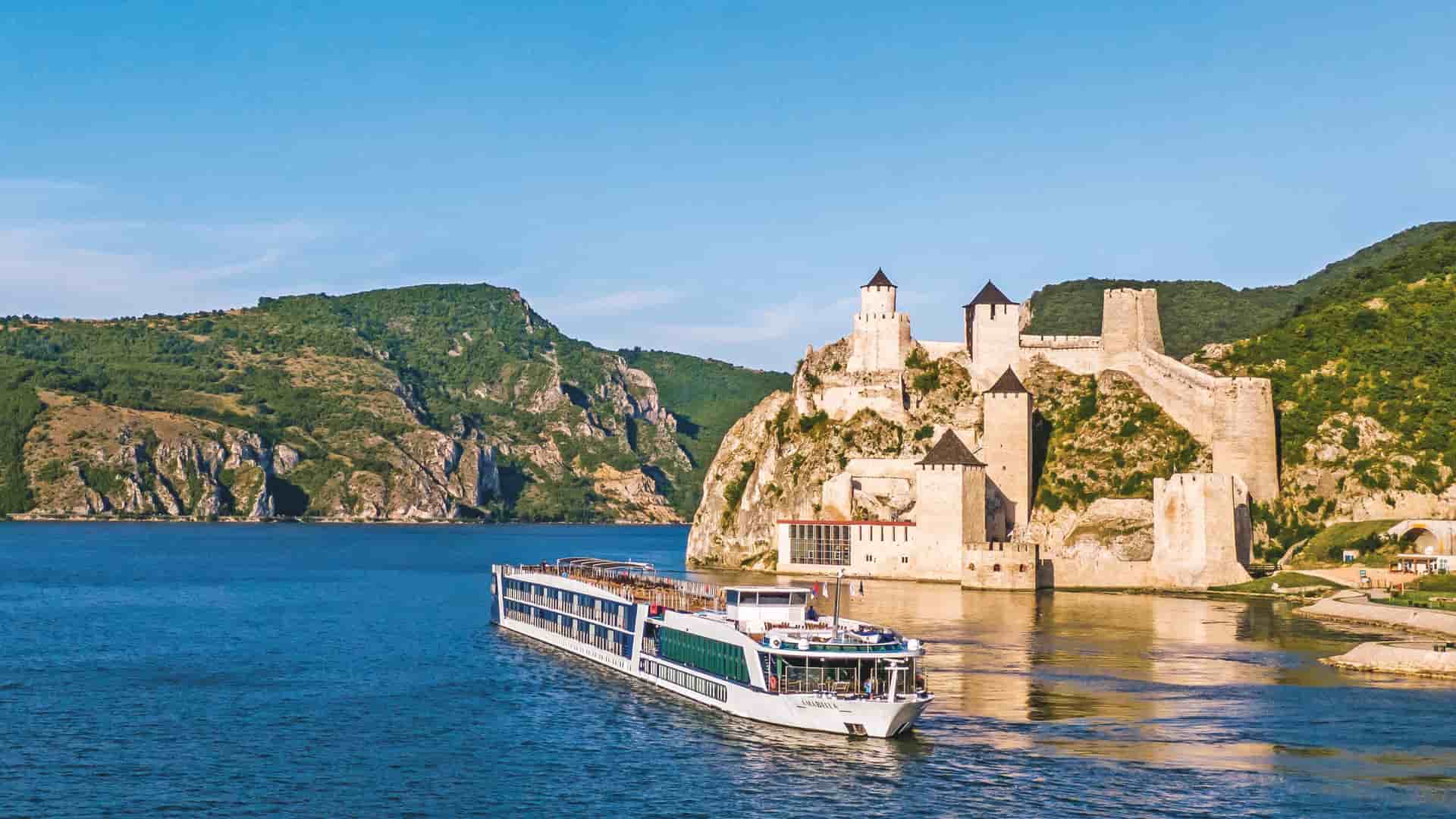   The AmaWaterways AmaBella river cruise ship sailing past the historic Golubac Fortress on the Danube River in Serbia, a medieval castle built into a cliffside.