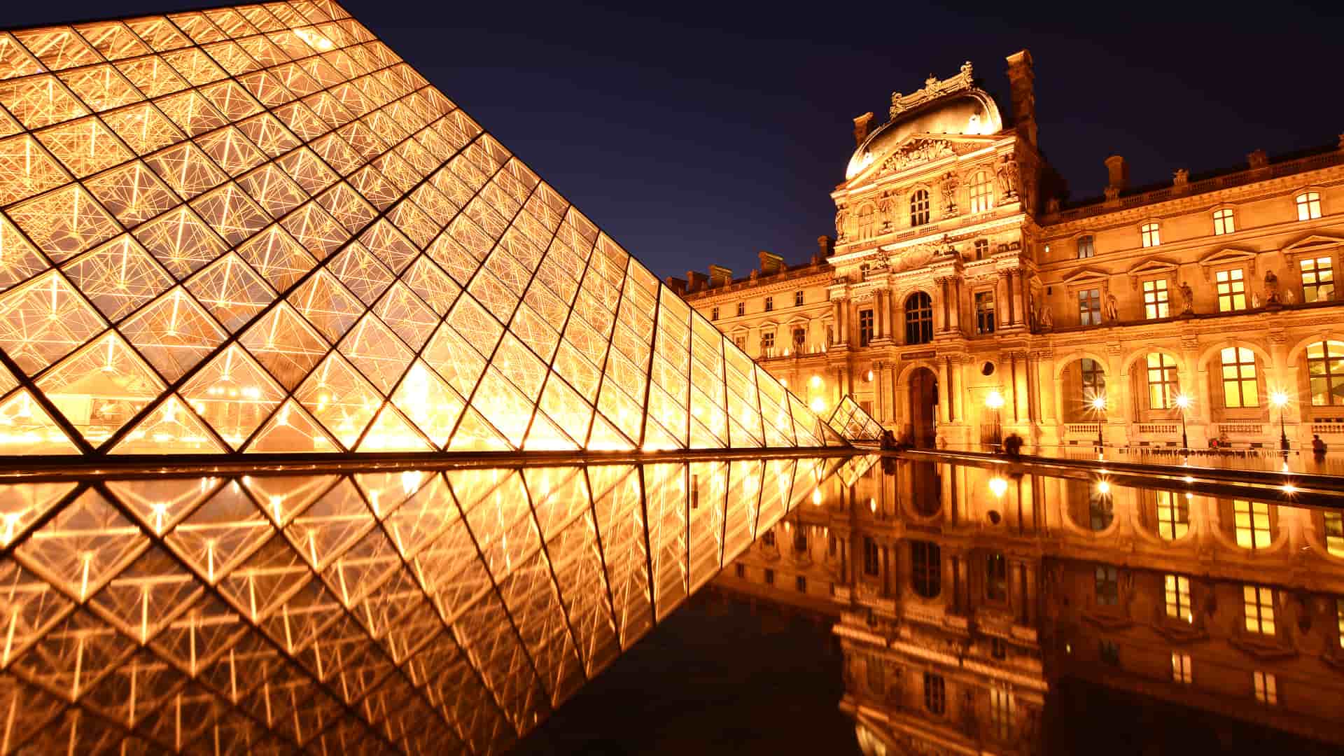 Night view of the illuminated Louvre Pyramid reflecting in the water, with the Louvre Palace in the background under a dark sky.