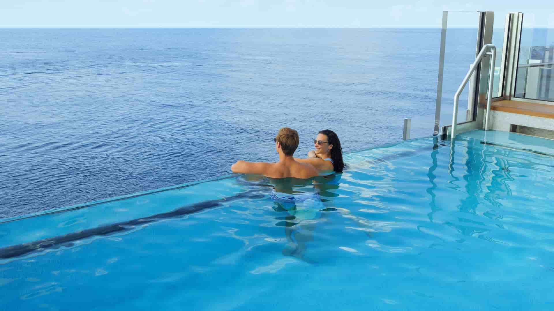 A couple is relaxing in the infinity pool on the Norwegian Cruise Line's Norwegian Luna ship, leaning on the edge and looking out at the vast ocean under a clear, blue sky.