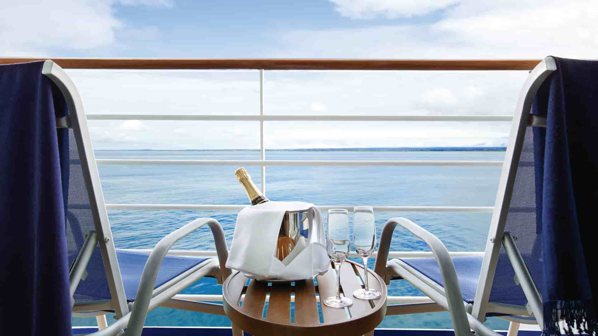 A bottle of champagne and 2 lounge chairs on the balcony of a penthouse suite overlooking a bright blue ocean on a beautiful day at sea on an Oceania cruise.