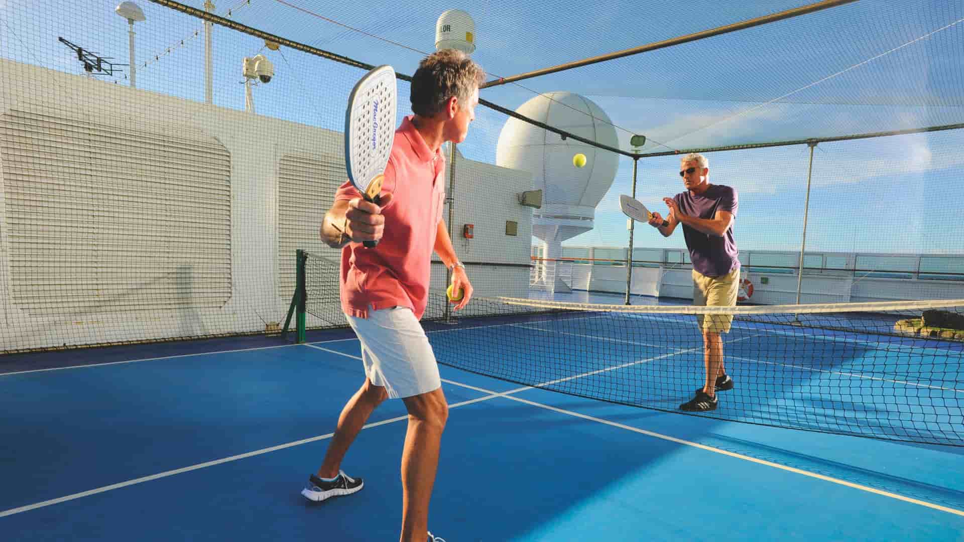 Two men on the Oceania Cruises' Sirena ship are playing pickleball on the top deck, with the man in the foreground wearing a pink shirt and white shorts, and the man in the background wearing a purple shirt and khaki shorts.