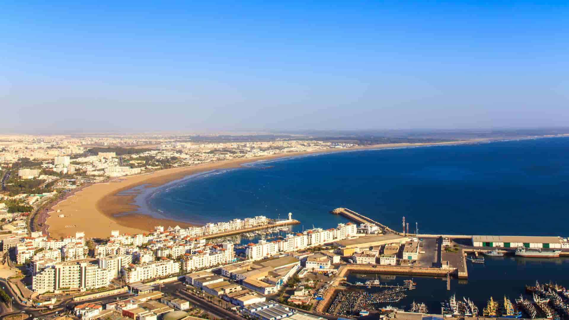 An aerial view of Agadir, Morocco, showcasing the city's long sandy beach and curving coastline, with a busy marina and port in the foreground.