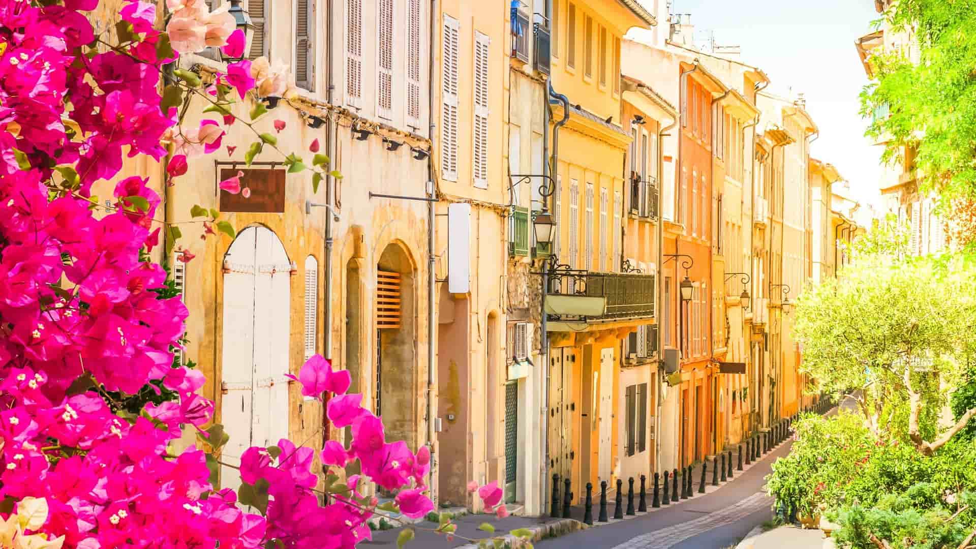 A picturesque street scene in Aix-en-Provence, France, with a row of historic pastel-colored buildings and vibrant pink bougainvillea flowers in the foreground.