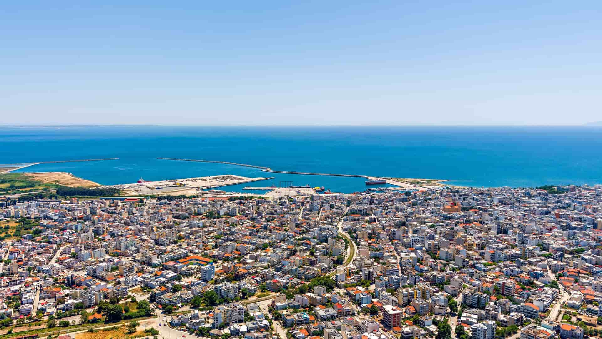 An aerial view of the coastal city of Alexandroupolis, Greece. Densely packed white buildings with red roofs extend to a large, busy port, where a long breakwater and piers jut into the deep blue Aegean Sea. The sky is clear and bright blue, stretching to meet the sea at the horizon.