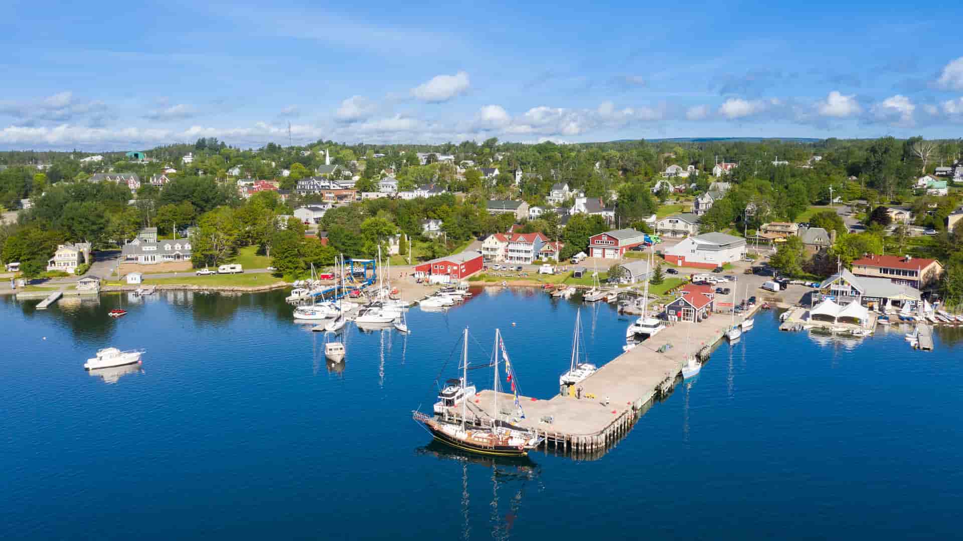 An aerial view of the picturesque waterfront in Baddeck, Nova Scotia, Canada. The deep blue water is filled with sailboats and motorboats docked at a long pier, which extends from the shoreline. A charming town with colorful houses and buildings is visible among the lush green trees, all under a bright blue sky with scattered white clouds.