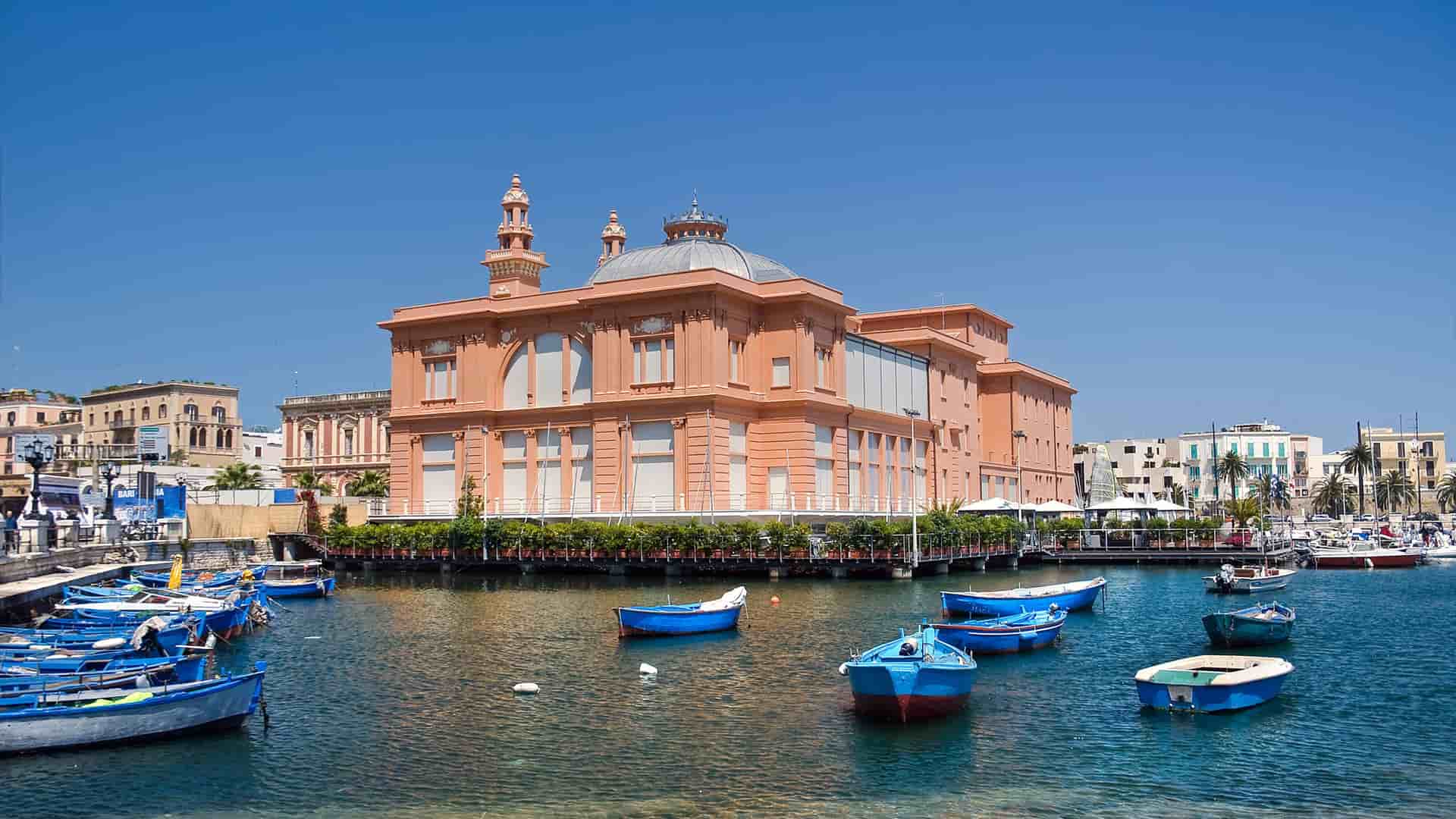 A scenic shot of the historic Teatro Petruzzelli in Bari, Italy, with its distinctive pink facade and a view of small, colorful fishing boats moored in the harbor.
