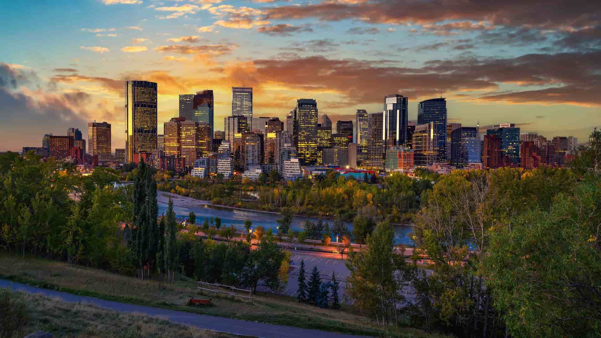 A panoramic, elevated view of the Calgary, Alberta skyline at sunset, with a collection of modern, illuminated skyscrapers rising behind the Bow River. The foreground is a lush, green park with trees and a walking path, all set under a dramatic sky with vibrant orange, yellow, and blue clouds.