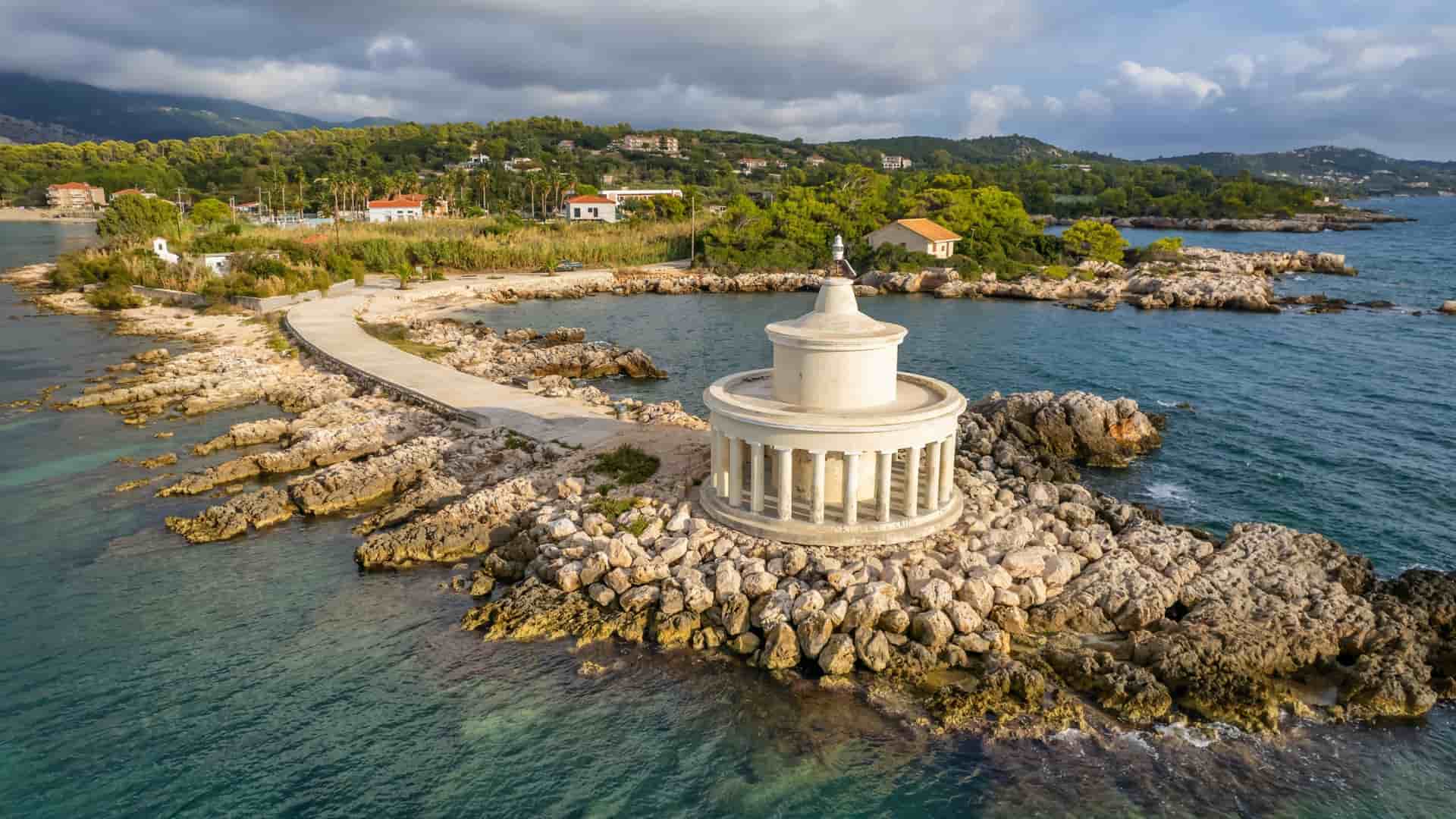 An aerial shot of the historic Fanari Lighthouse on a small rocky islet, connected by a causeway to the mainland of Argostoli, Cephalonia, with villas and lush hills in the background.