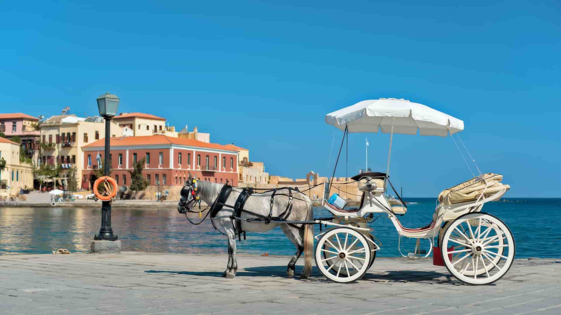 A picturesque scene of a horse-drawn carriage on the waterfront of Chania, Crete, with historic Venetian harbor buildings and the deep blue Mediterranean Sea in the background.