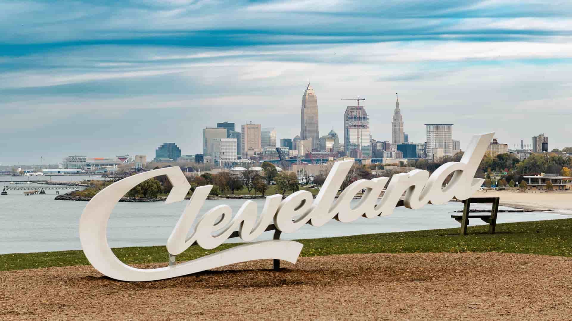 A panoramic shot of the Cleveland skyline, with its downtown skyscrapers and Lake Erie in the background, featuring the large white "Cleveland" sign in the foreground.