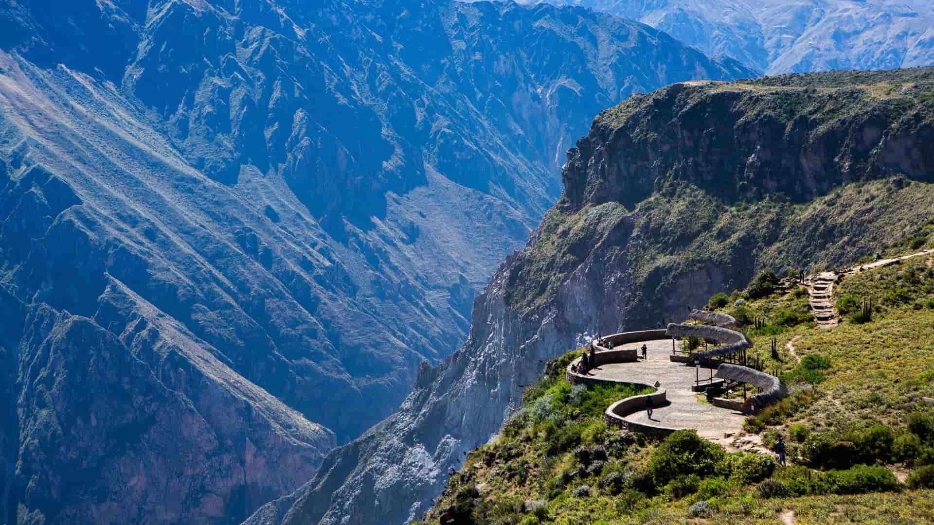 A stunning aerial view of the deep Colca Canyon in Peru, with tourists standing on a winding observation deck overlooking the dramatic, steep mountain slopes.
