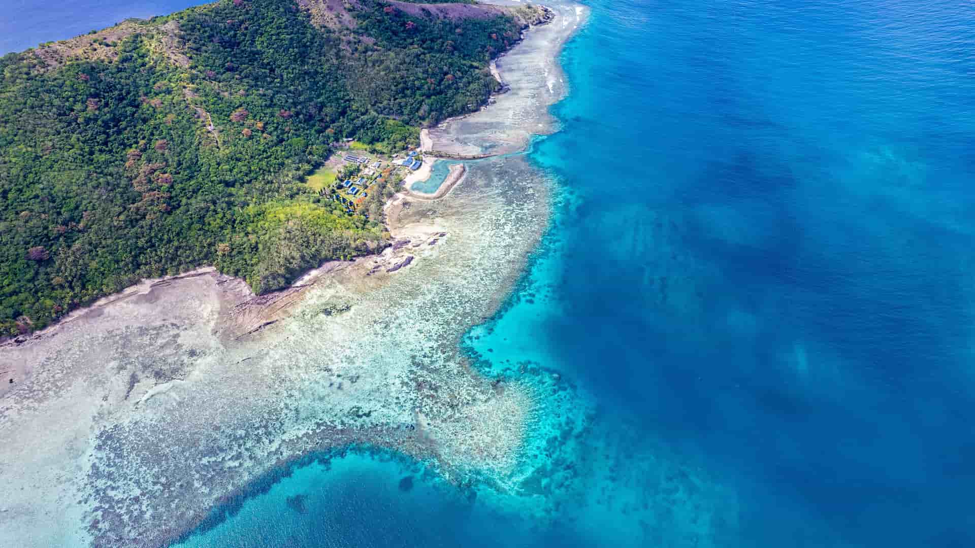 "An aerial view of the stunning Coral Coast in Fiji, showing a lush green island meeting the vibrant blue water of the Pacific Ocean, with a shallow coral reef visible beneath the surface.  "