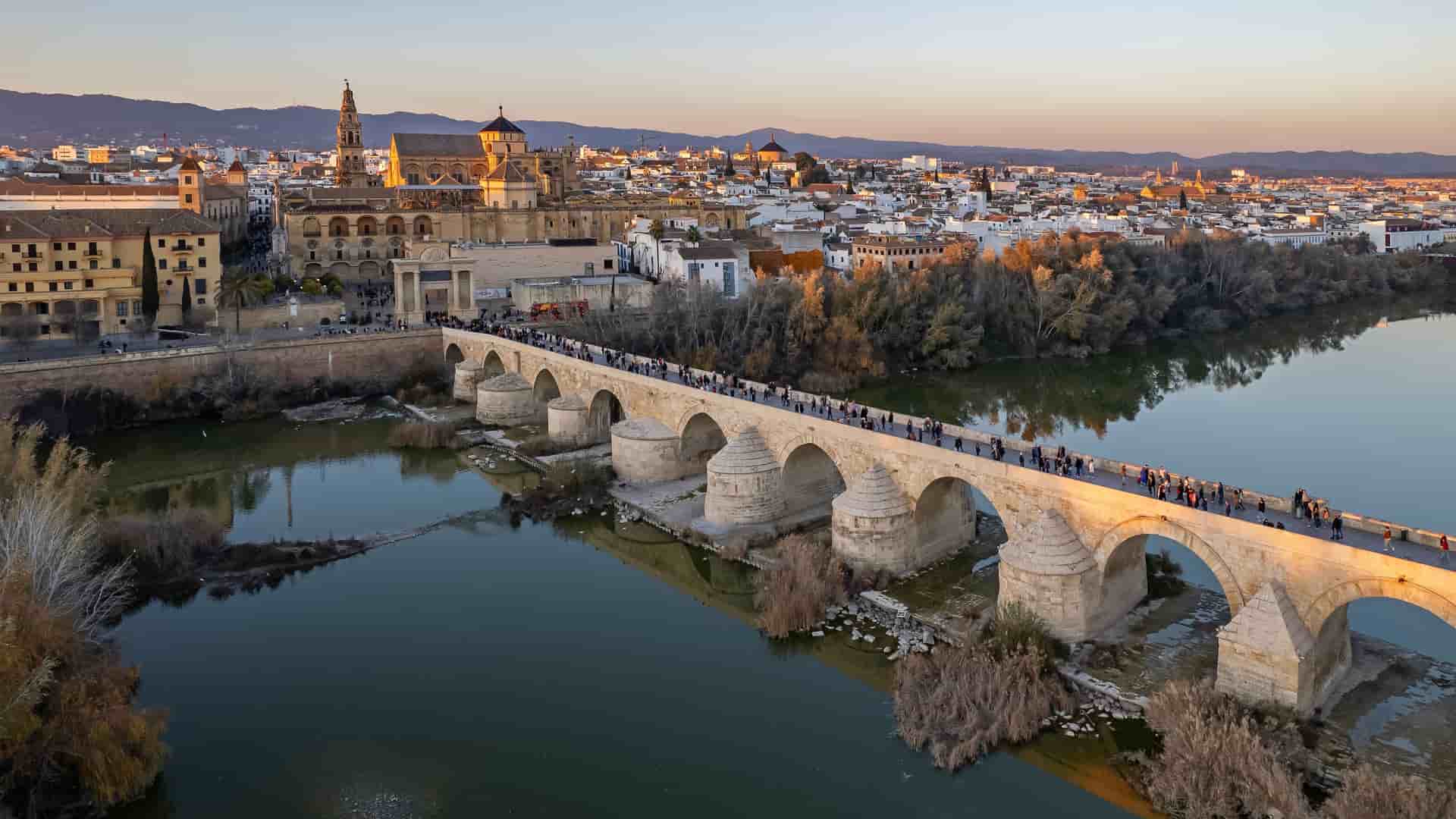 "An aerial view of the historic Roman Bridge of Córdoba, Spain, with tourists walking across its many arches over the Guadalquivir River, leading to the ancient Mezquita-Cathedral and the city skyline during sunset.  "