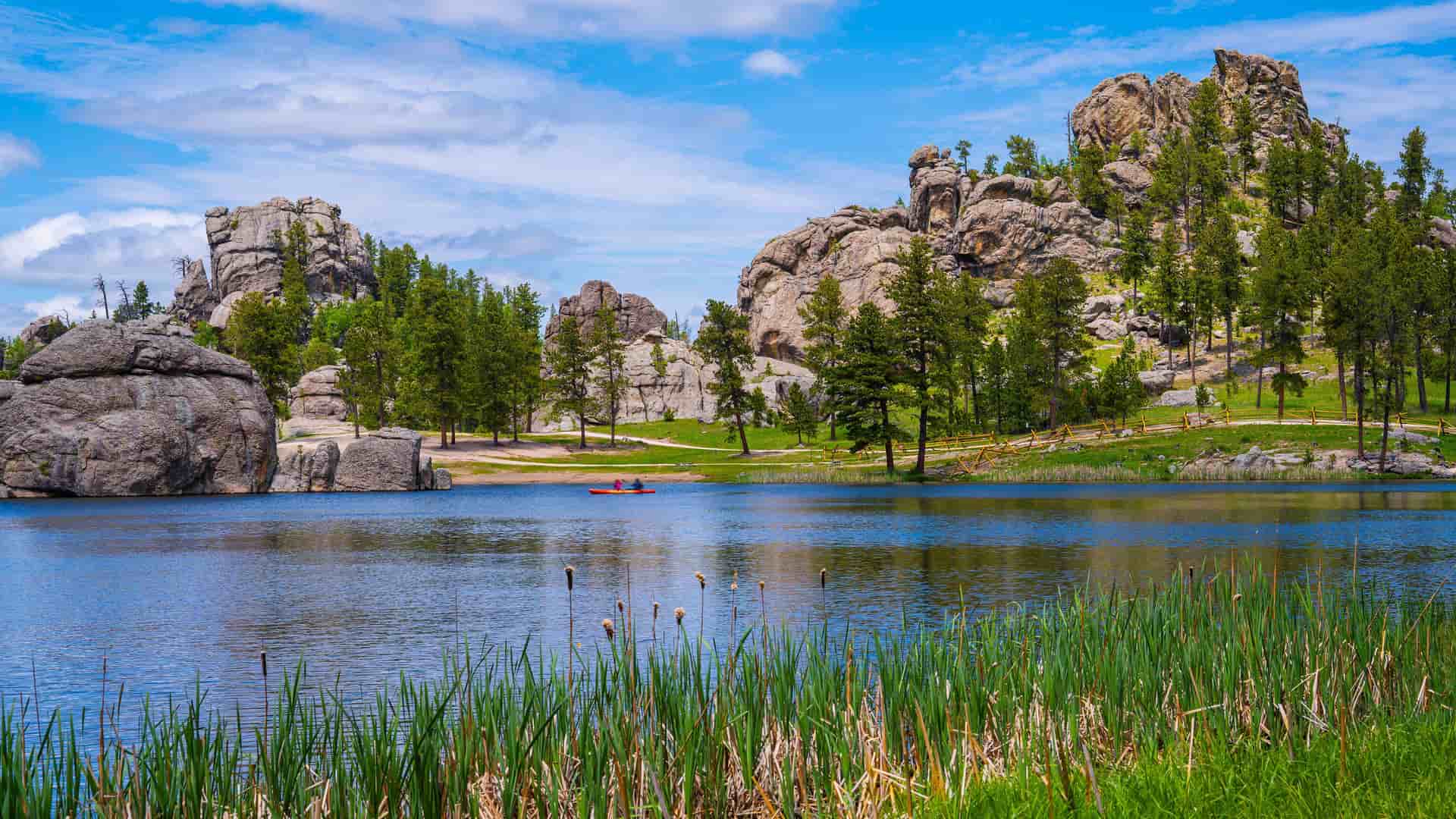 "A tranquil lake with lush reeds and grasses in the foreground, reflecting the surrounding granite rock formations and ponderosa pine trees, with two people in a red kayak on the water in Custer State Park, South Dakota.  "