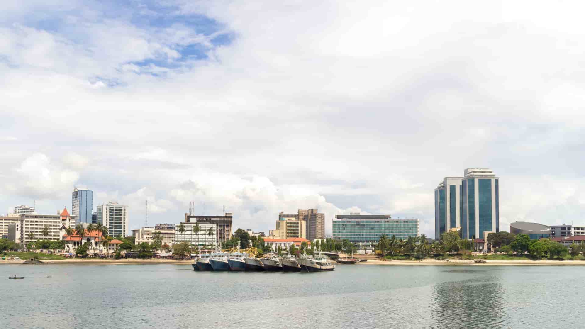 A view of the Dar es Salaam skyline in Tanzania from the water, showing modern office buildings behind traditional wooden boats moored in the harbor.