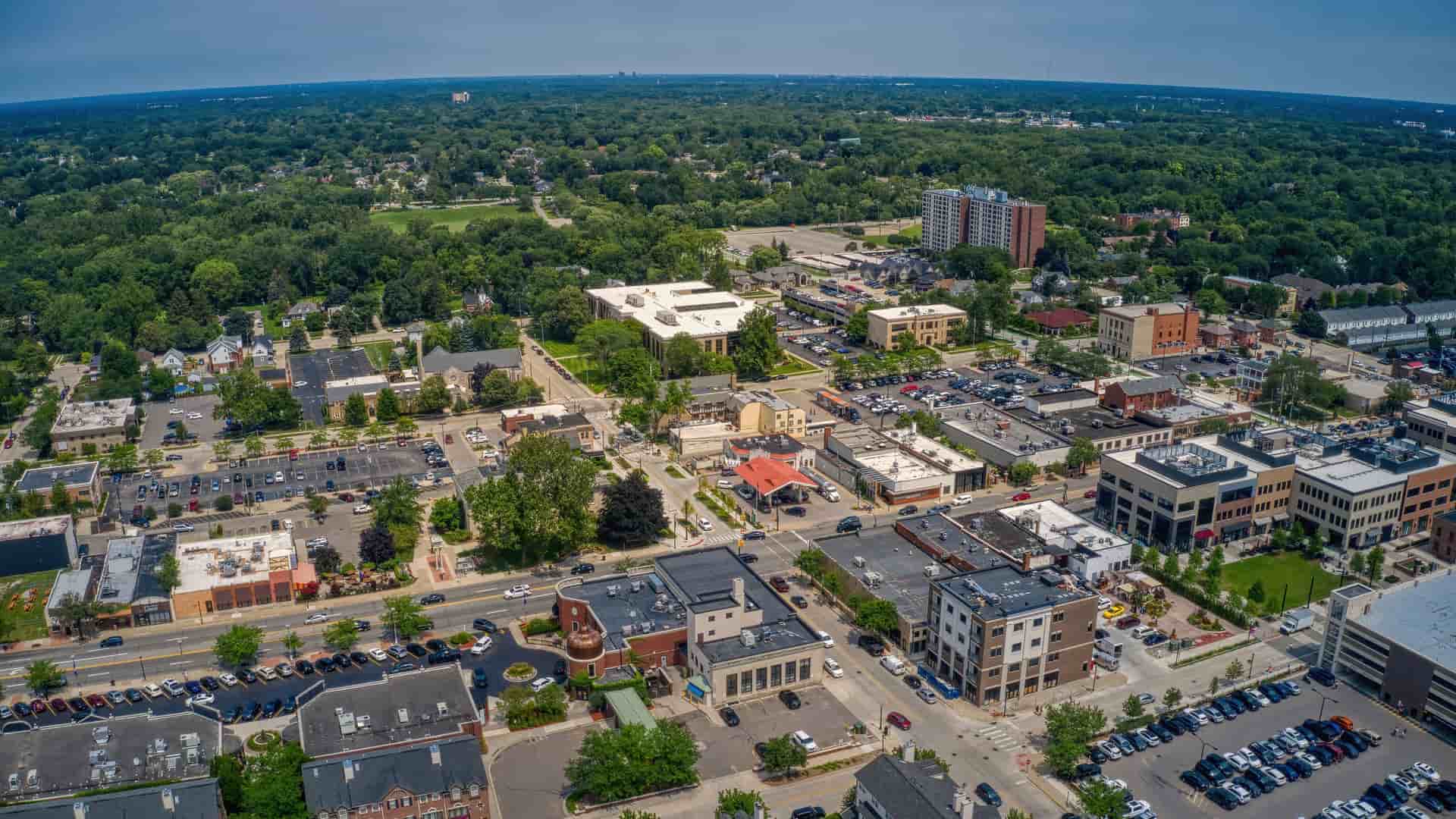 "An aerial view of downtown Dearborn, Michigan, showcasing a mix of modern and historic buildings, commercial areas, and residential neighborhoods surrounded by the dense green trees of the city.  "