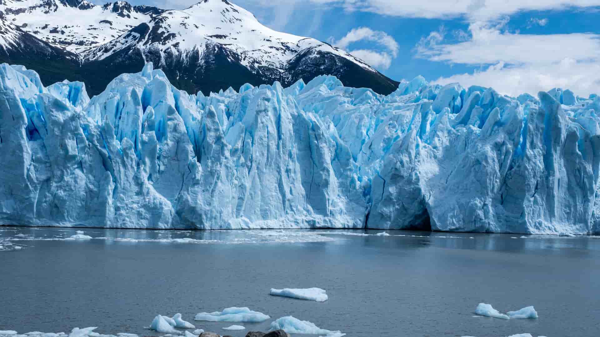 "A magnificent view of the bright blue and white face of the Perito Moreno Glacier in Patagonia, Argentina, with small icebergs floating in the frigid lake and snow-capped mountains in the background.  "