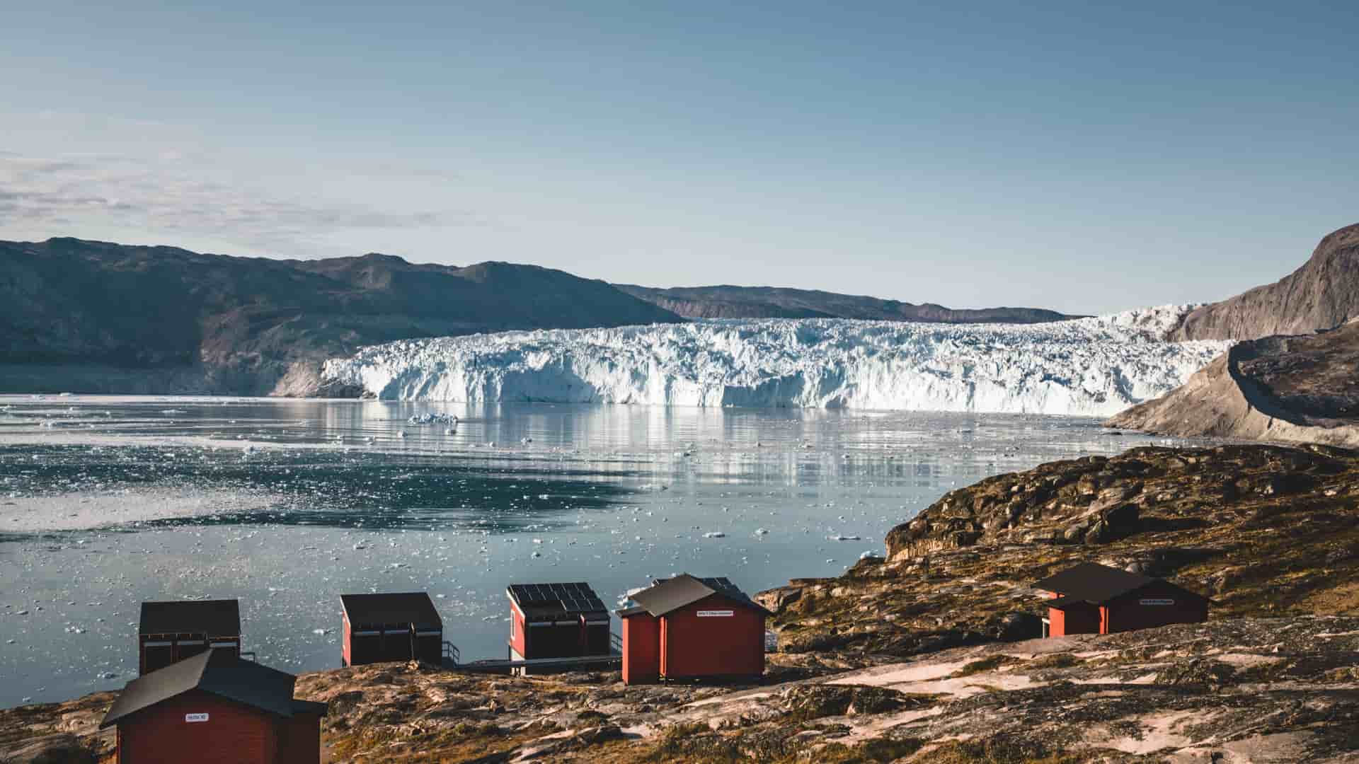 A panoramic view of the massive Eqip Sermia Glacier in Greenland, with a cluster of red huts on the rocky shore and ice chunks floating in the water under a clear blue sky.