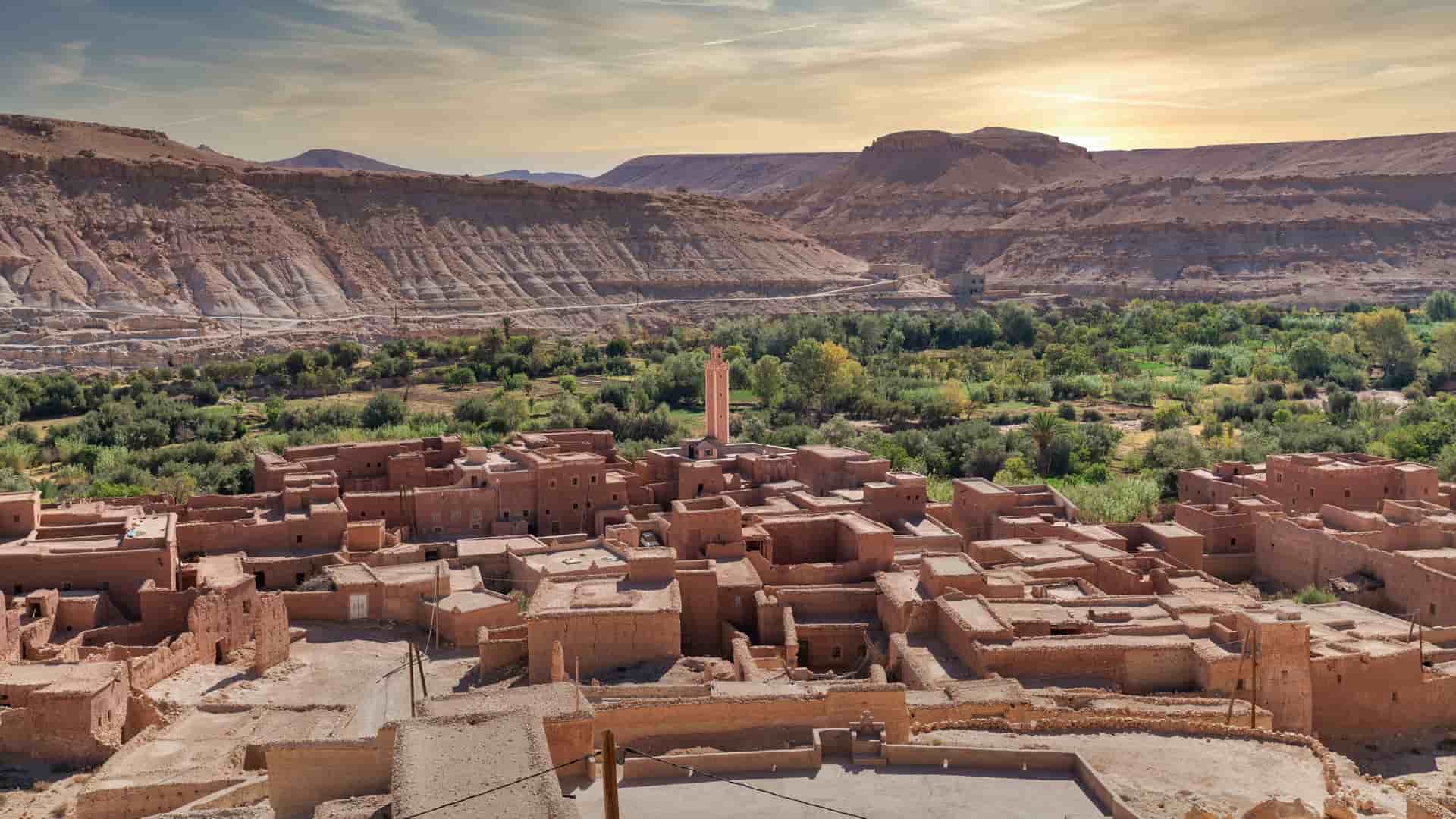 "A high-angle view of a traditional clay village in Erfoud, Morocco, with a single minaret in the center and a lush green oasis of date palms and other trees in a valley, surrounded by the barren, rocky landscape of the Atlas Mountains.  "