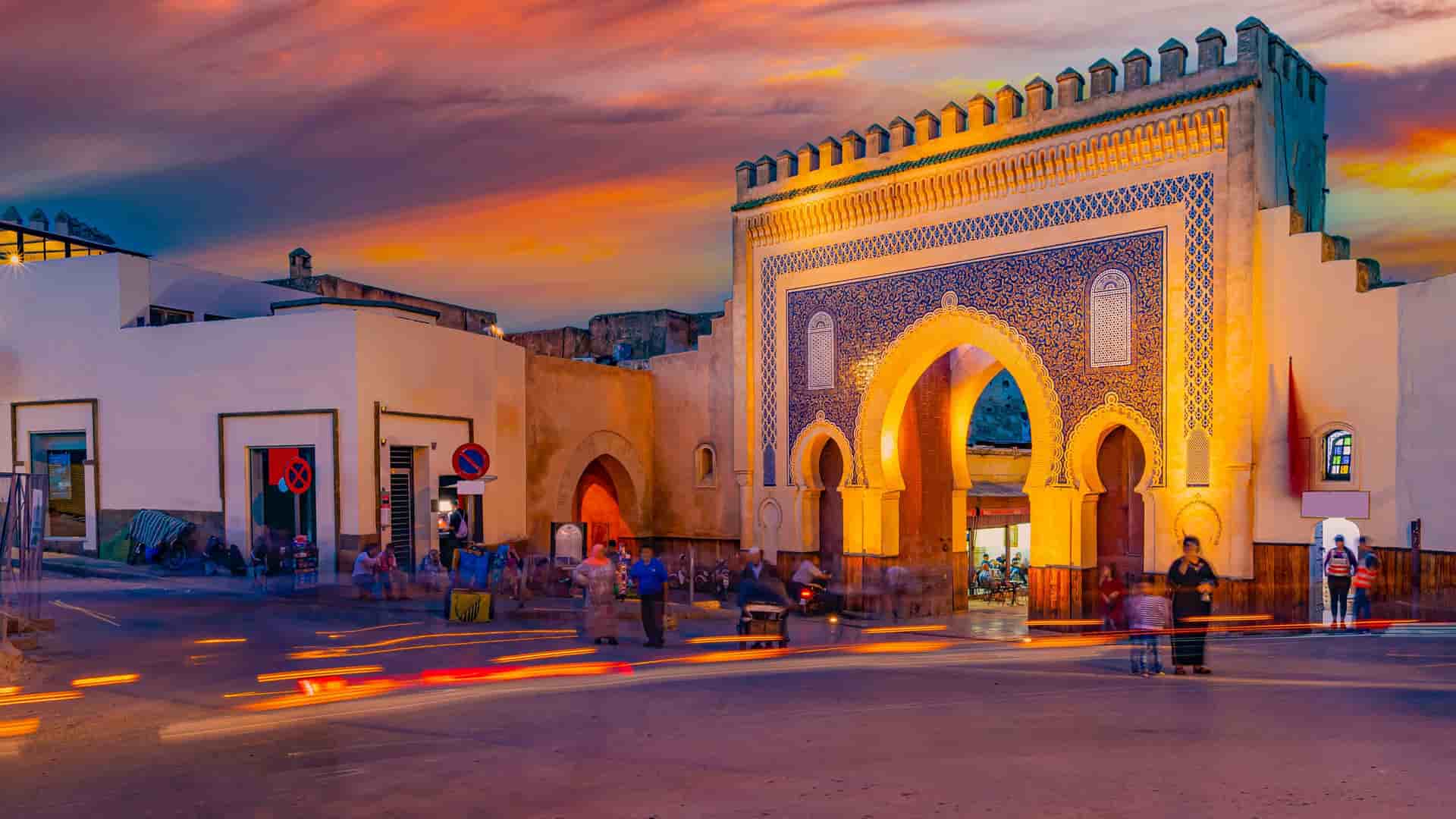 "A beautiful sunset view of the iconic Bab Bou Jeloud gate in Fez, Morocco, with its intricate blue and green tile mosaics, glowing in the light as people bustle through the archway.  "