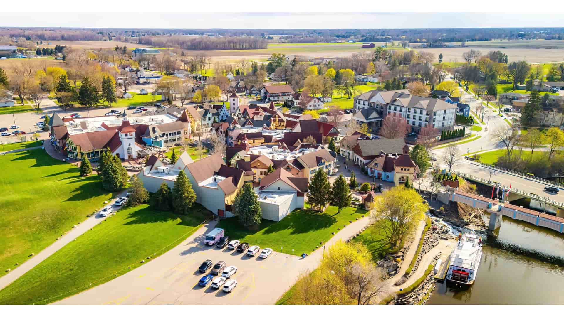 "An aerial view of the Bavarian-style town of Frankenmuth, Michigan, showing its unique architecture, a riverboat on the Cass River, and surrounding green fields.  "