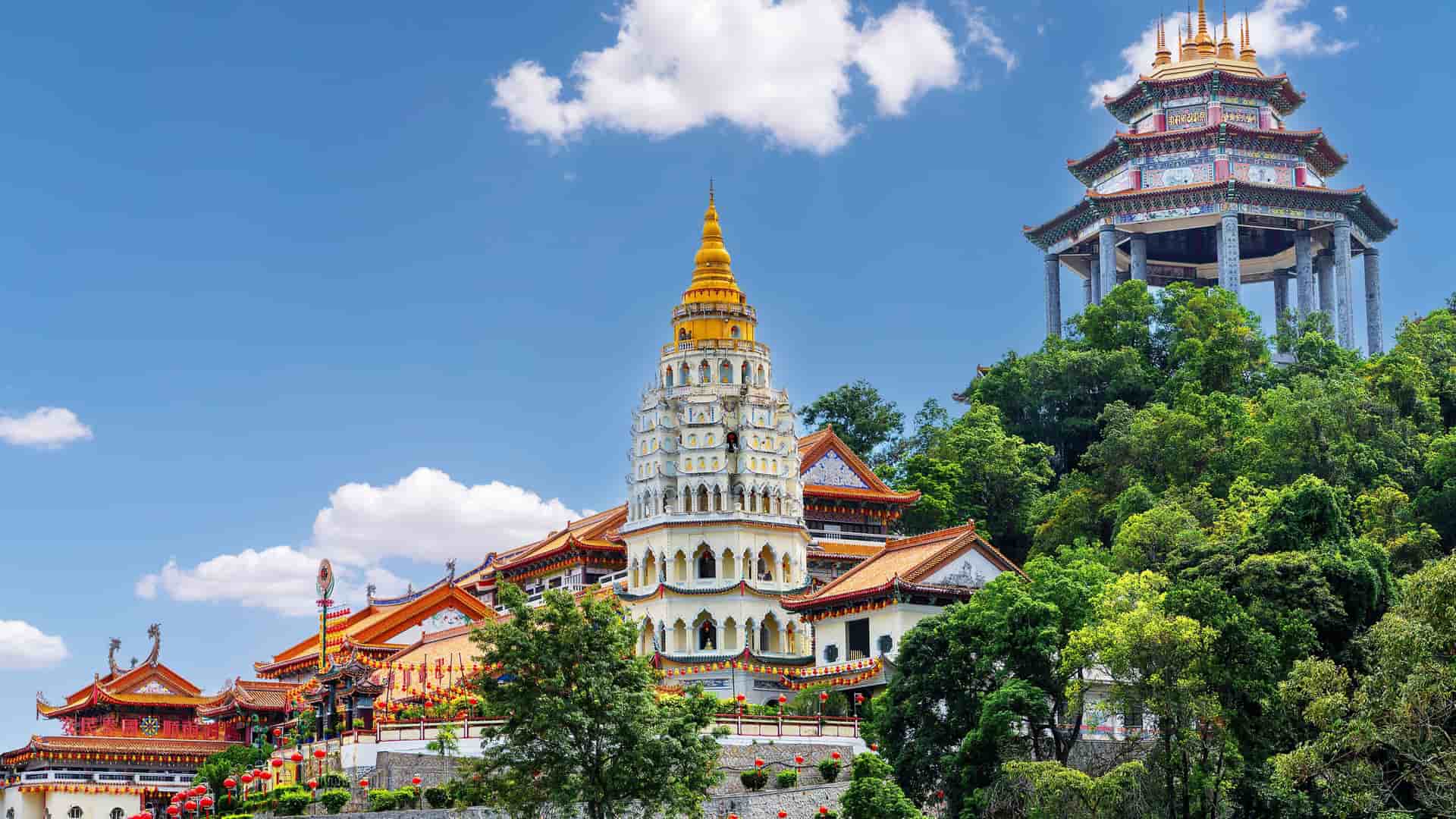A vibrant wide-angle shot of the Kek Lok Si Temple in Penang, Malaysia, showcasing its colorful pagodas, lush trees, and intricate architecture against a bright blue sky.