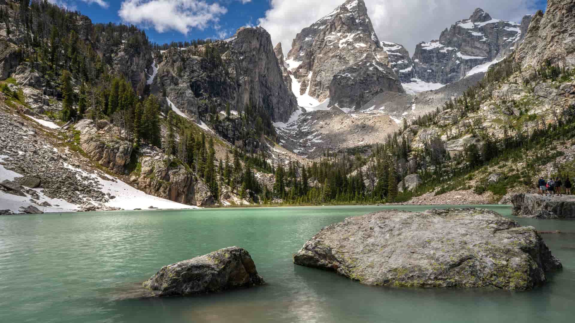 "A beautiful landscape view of Grand Teton National Park, featuring the turquoise waters of Taggart Lake with large boulders in the foreground and the jagged, snow-dusted mountains of the Teton Range in the background.  "