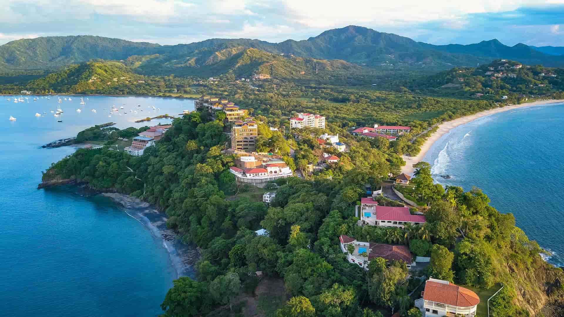 "An aerial view of the coastline of Guanacaste, Costa Rica, showing a verdant peninsula with luxury resorts and homes, with a white sand beach and boats moored in the clear blue water.  "