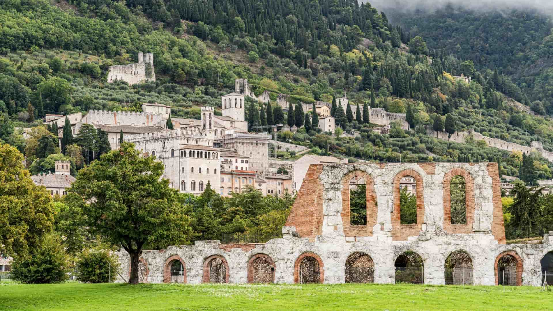 Ruins of a Roman archway and red brick structure in the foreground, with the medieval hill town of Gubbio, Umbria, Italy, visible on the slope of Mount Ingino behind a grassy field.