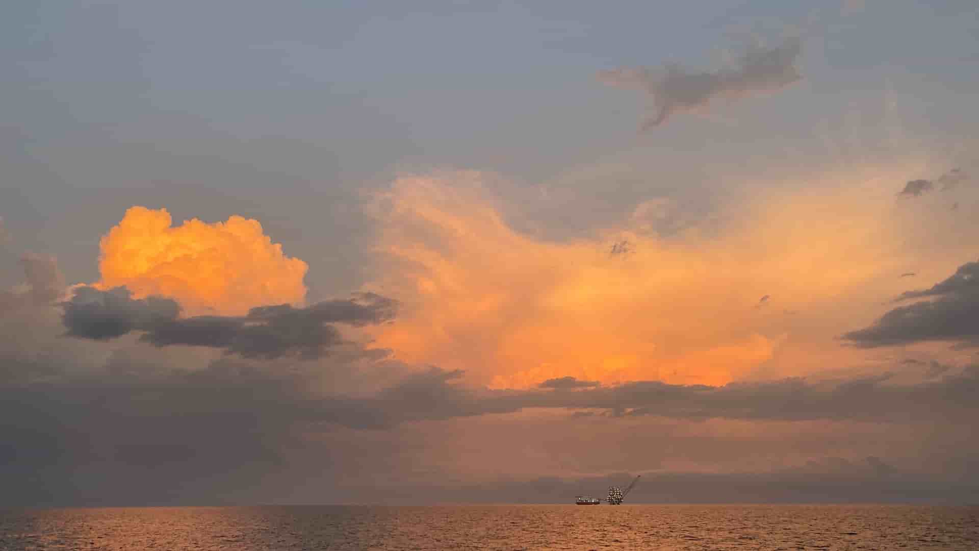A beautiful, dramatic sunset over the Gulf of Guinea, with the sun's warm light illuminating large clouds and a distant oil platform on the calm sea.