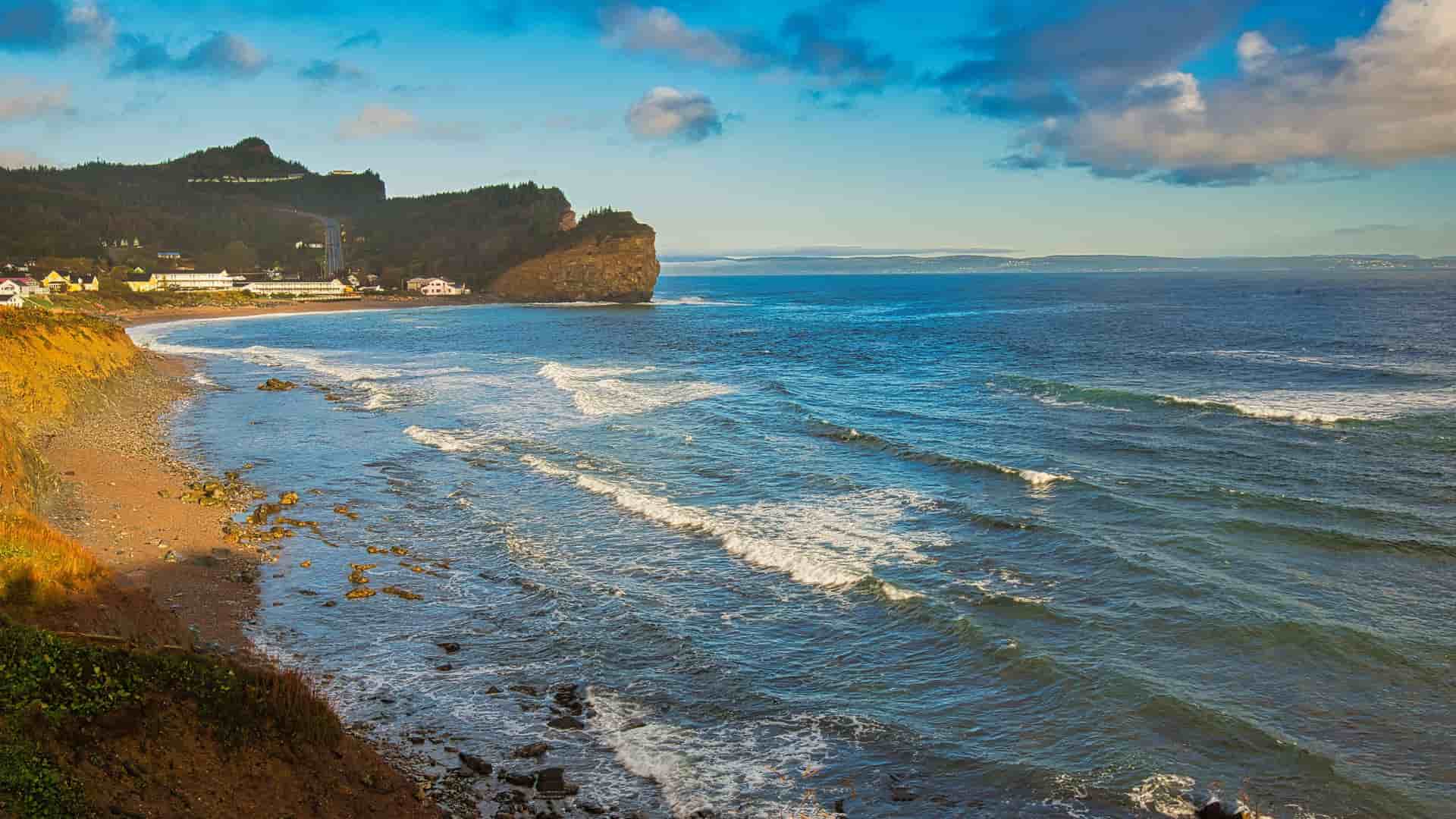 A panoramic view of the rugged coastline of the Gulf of St. Lawrence, with a sandy beach and small houses near a massive rock formation.