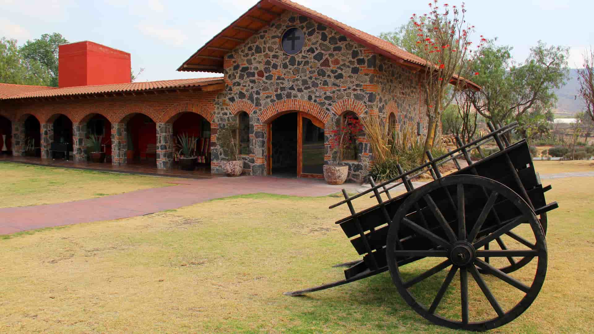 "A historic colonial-era building with a stone facade and red-tiled roof, part of the Hacienda Concepción in Peru, with a large, old wooden cart on the grassy lawn in front.  "