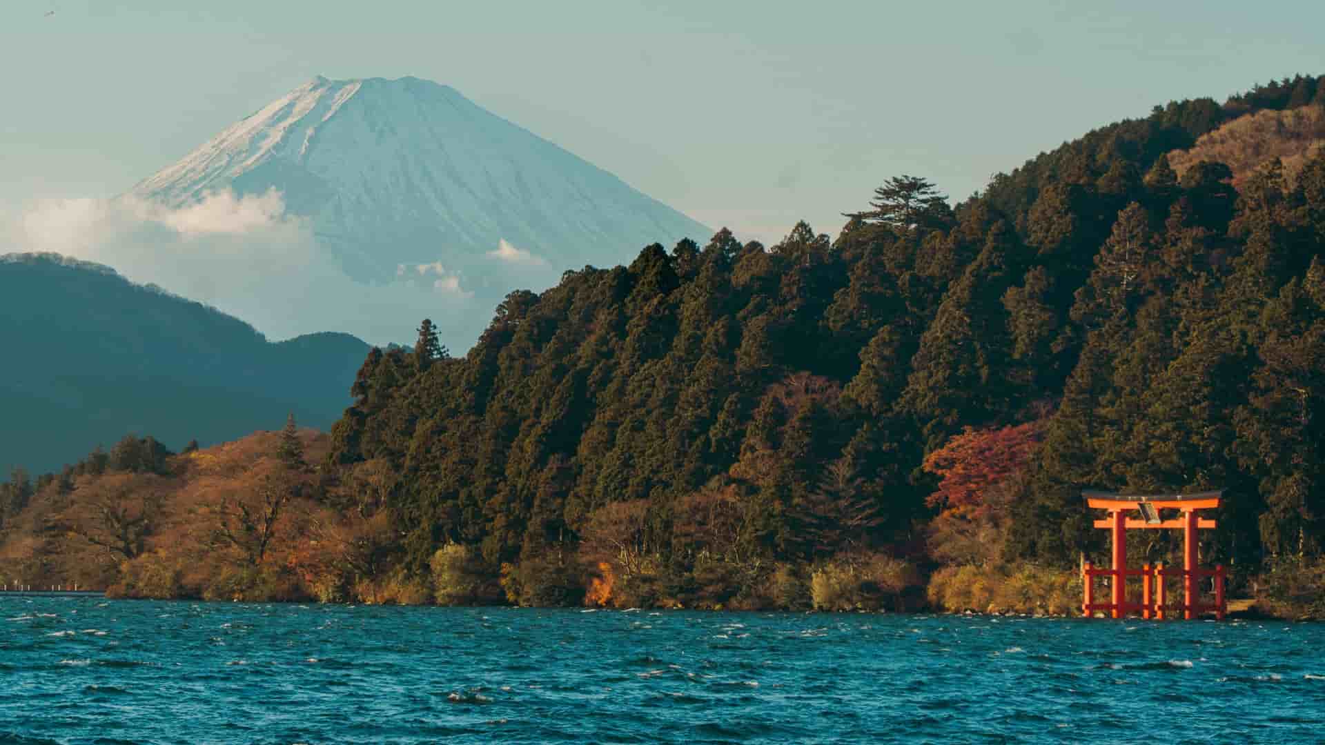 A scenic view across Lake Ashi in Hakone, Japan, with the iconic snow-capped peak of Mount Fuji visible in the background over a dense, pine-covered hillside, and a bright orange torii gate standing at the water's edge.