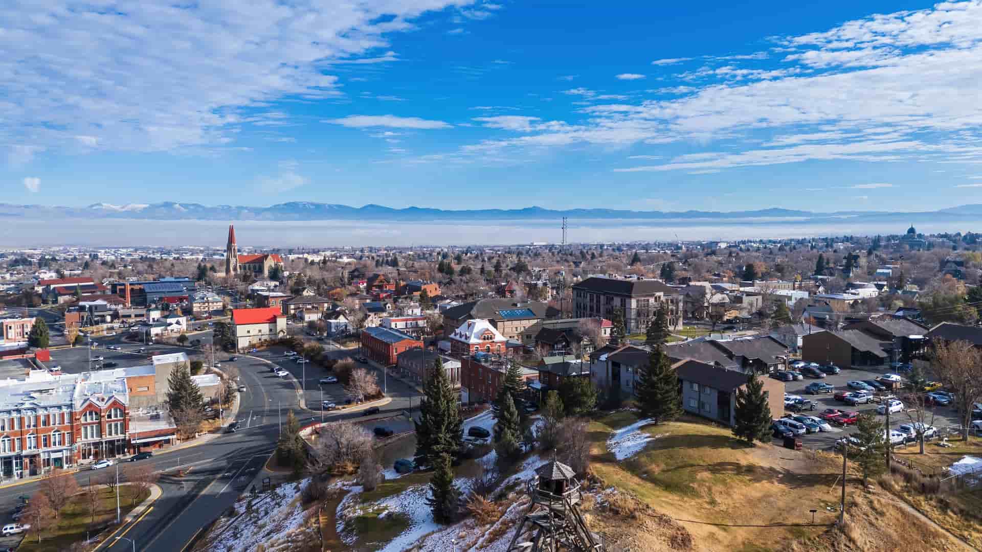"An aerial view of Helena, Montana, with the city's historic buildings and the iconic Cathedral of St. Helena in the foreground, and the snow-capped mountains of the Rocky Mountains in the distance under a blue sky.  "