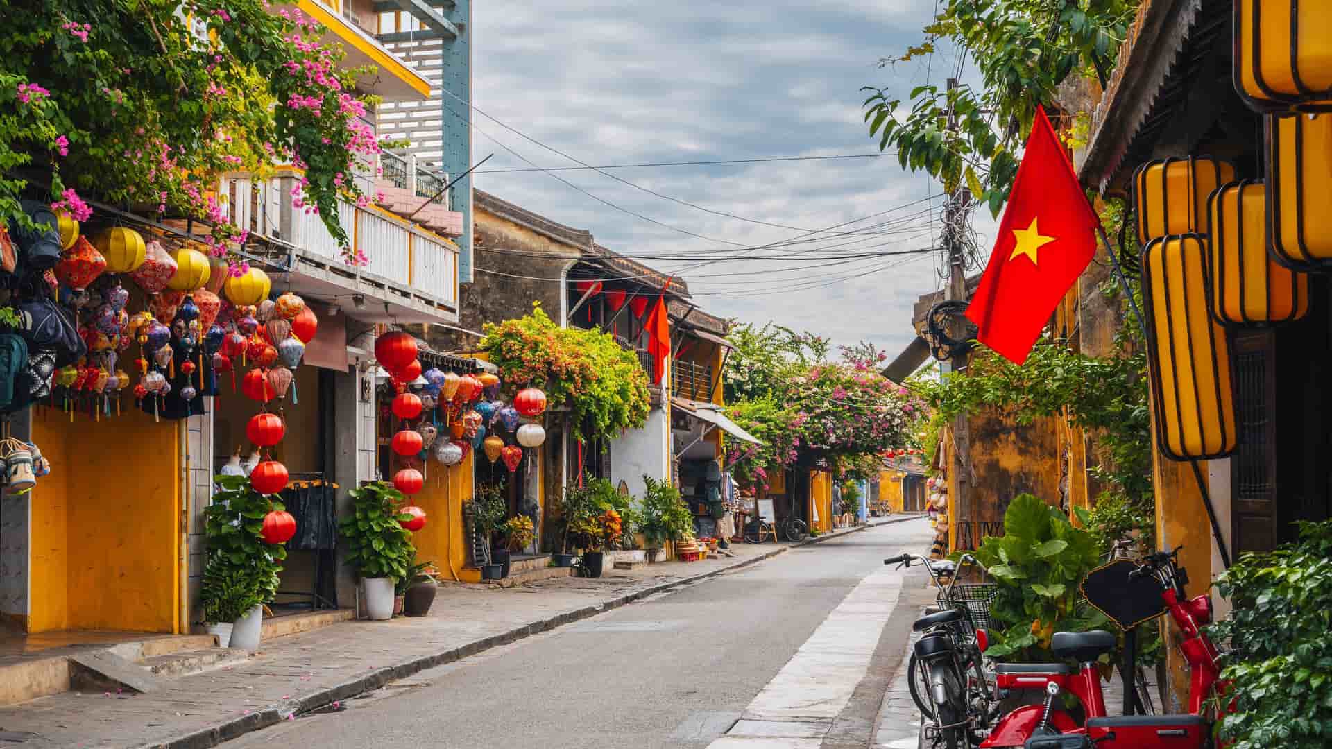 "A beautiful street in Hoi An, Vietnam, is lined with traditional yellow buildings adorned with colorful silk lanterns and blooming bougainvillea, with the Vietnamese flag flying from a storefront.  "