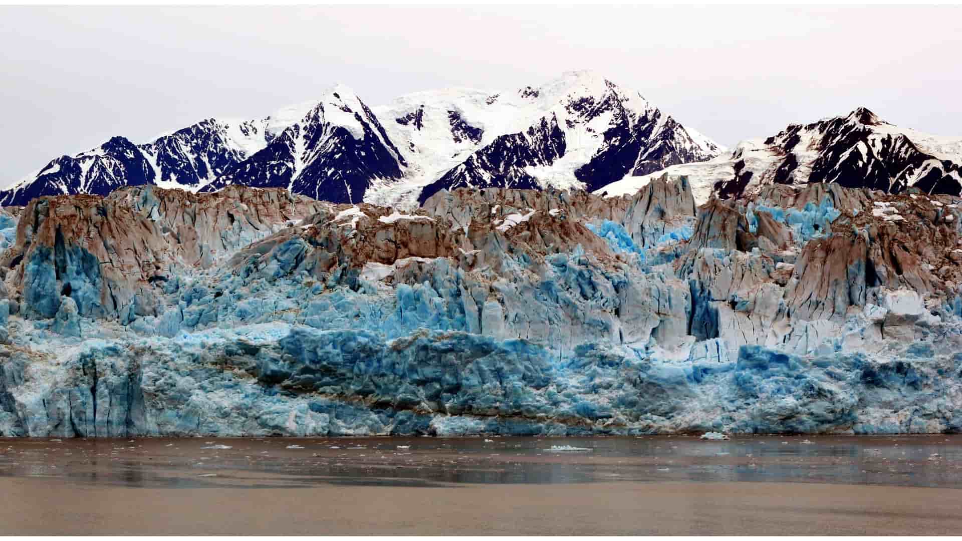 A stunning close-up of the Hubbard Glacier in Alaska, showcasing its immense, jagged face with beautiful blue and white ice against a backdrop of snow-capped mountains.