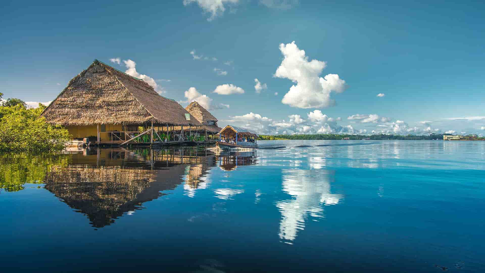 A wide-angle landscape shot of a tranquil lake in Iquitos, Peru, with reflections of thatched-roof houses and puffy clouds on the water.