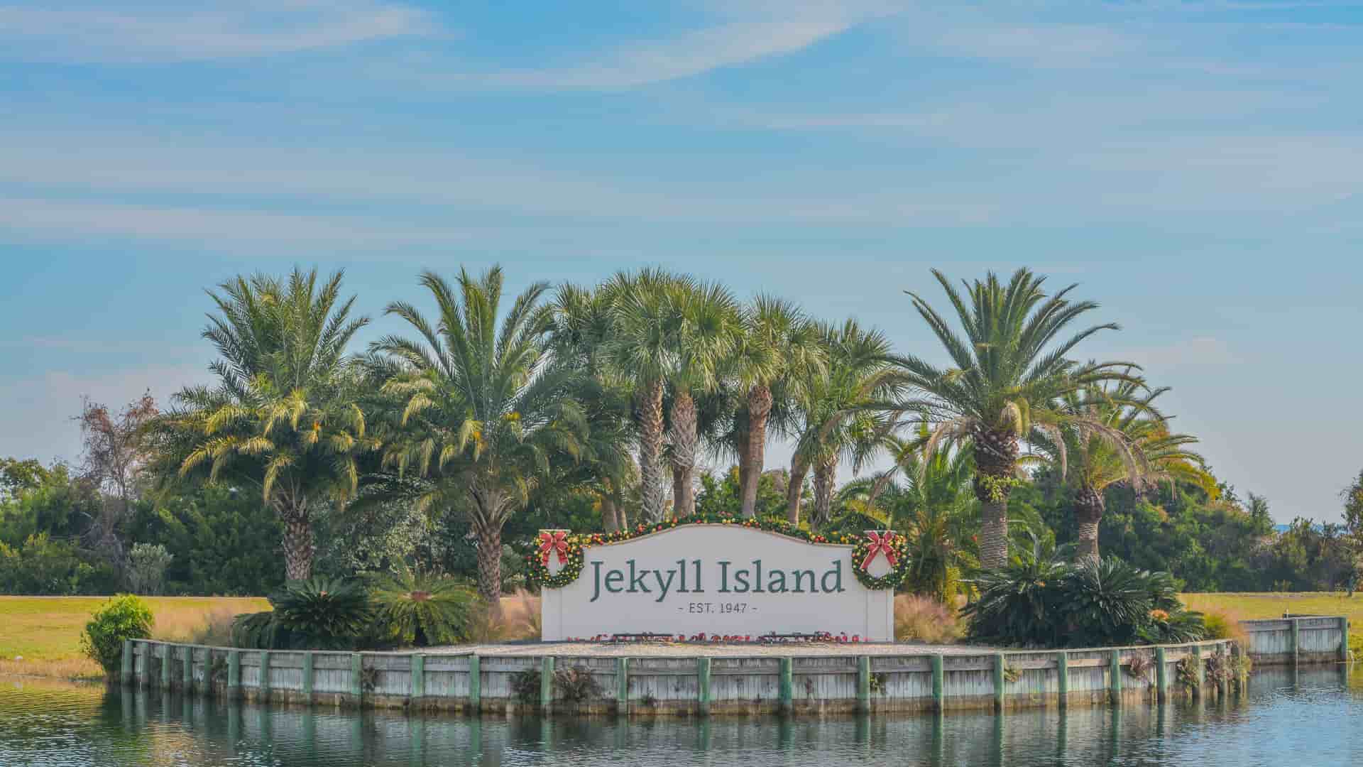 "A welcome sign for Jekyll Island, Georgia, with Christmas decorations on the front, surrounded by lush palm trees and water in the foreground.  "