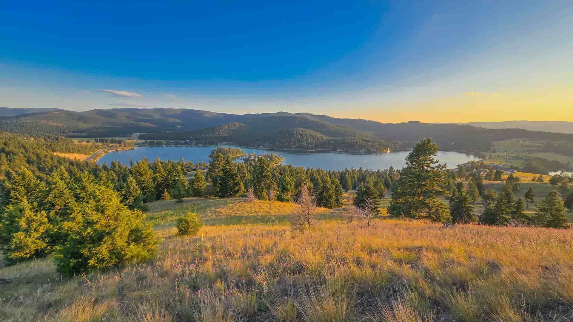"A panoramic sunset view over Flathead Lake, the largest freshwater lake in the western United States, with forested hills and a golden meadow in the foreground near Kalispell, Montana.  "