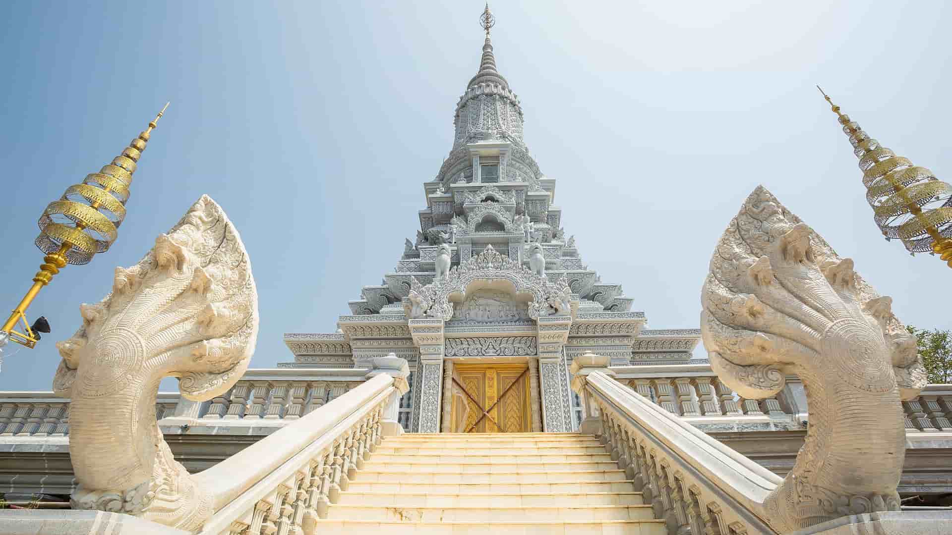 A stunning low-angle view of the ornate Udong mountain pagoda in Kampong Tralach, Cambodia, featuring a grand staircase flanked by guardian Naga snake statues leading up to the intricately decorated temple.