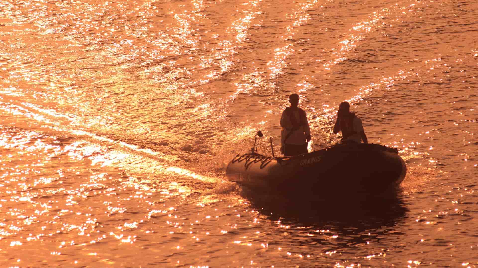 Two people ride a small rubber boat across the glittering, orange-colored water at sunset near Kenanga Village, Indonesia.