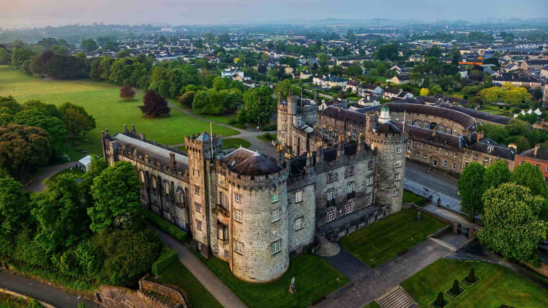 An aerial view of the historic Kilkenny Castle in Kilkenny, Ireland, featuring a large, medieval stone structure with rounded corner towers and manicured green lawns, set against the backdrop of the sprawling, low-rise town extending into the hazy distance.