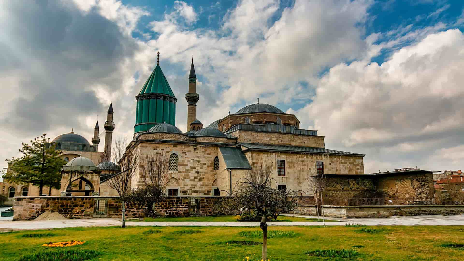 "A panoramic shot of the historic Mevlana Museum in Konya, Turkey, with its famous fluted green dome and multiple minarets under a dramatic cloudy sky, with a green grassy lawn in the foreground.  "