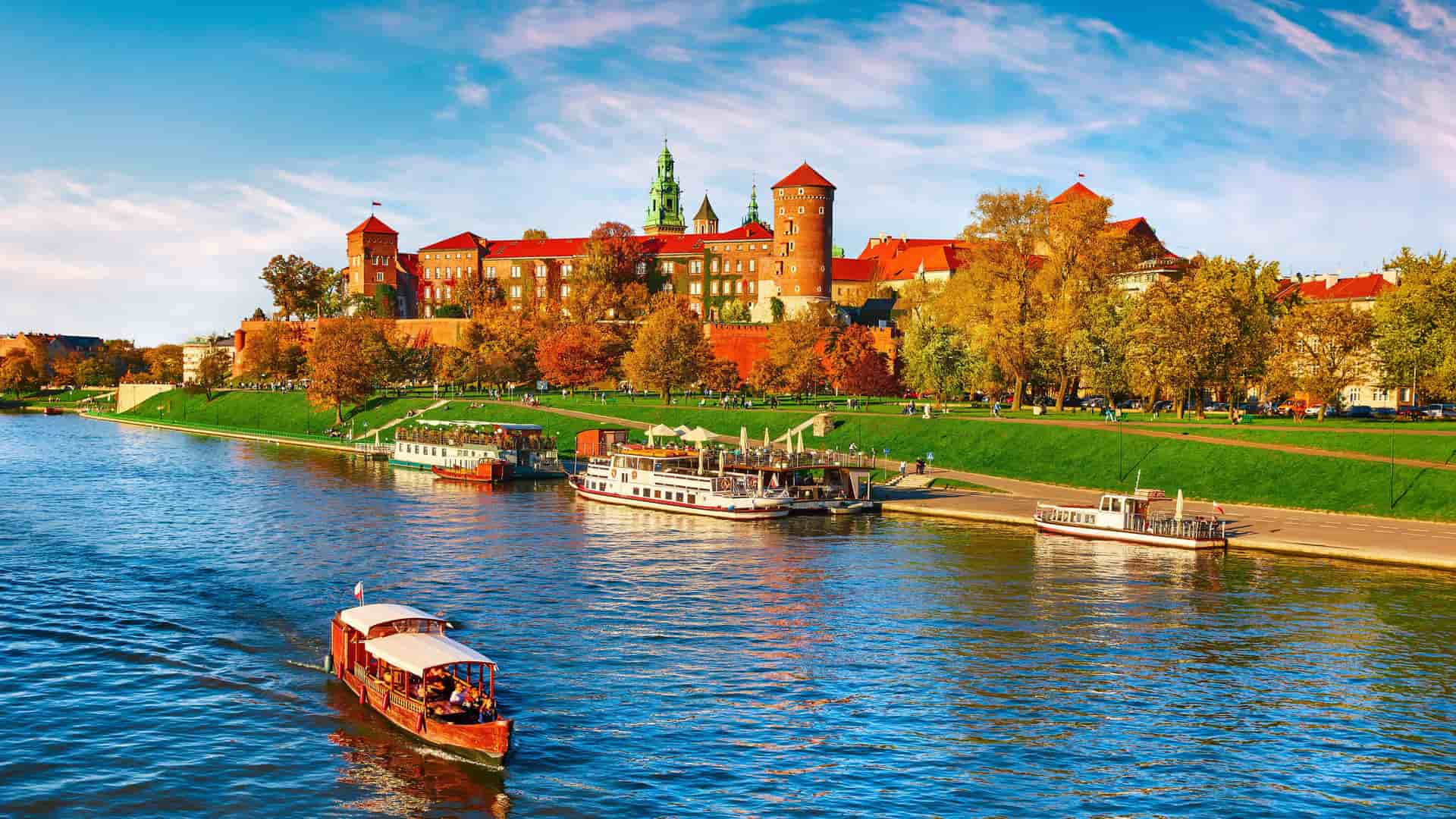 "A vibrant autumn view of Wawel Royal Castle in Krakow, Poland, with its red brick walls and towers, situated on a hill overlooking the Vistula River with several tour boats.  "