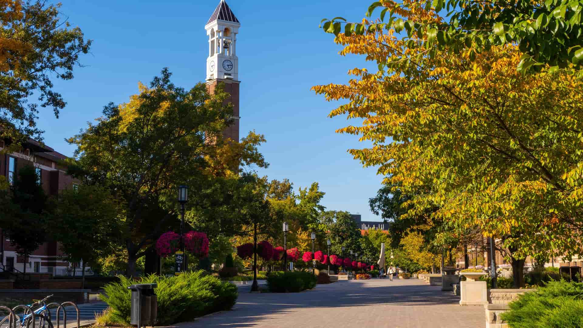 "A vibrant autumn scene on the campus of Purdue University in Lafayette, Indiana, showing the iconic Bell Tower and a path lined with trees in full fall foliage.  "