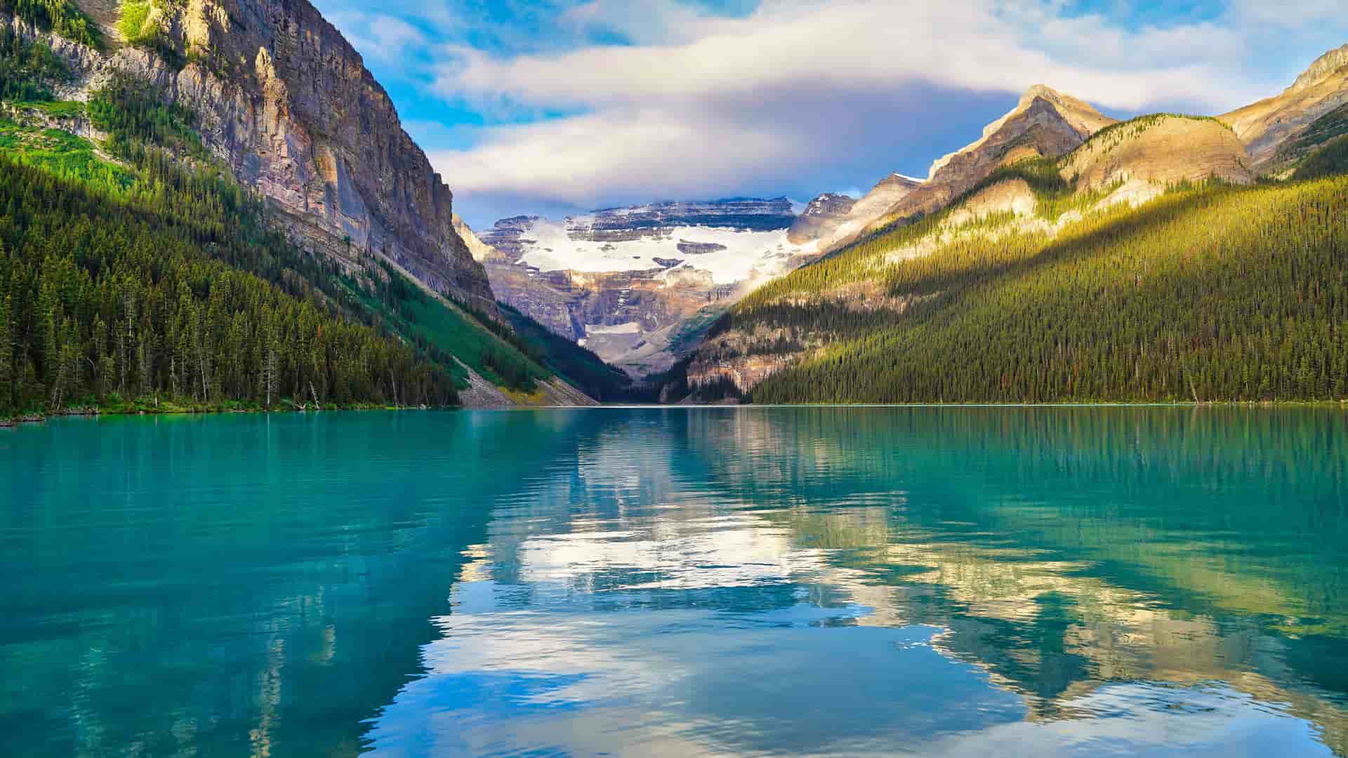 "A picturesque view of the serene turquoise waters of Lake Louise, surrounded by towering, forested mountains and the snow-capped Mount Victoria in the distance in Banff National Park, Canada.  "